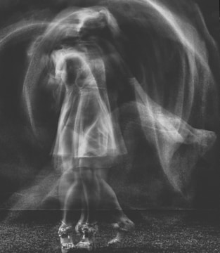 A blurred, monochrome image of a person in motion, captured in a way that creates a ghostly or ethereal effect. The figure appears to be spinning or dancing, with a dress and shoes faintly visible.