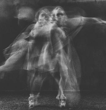 A blurred, ghostly figure in motion, captured in black and white, creating an ethereal and fluid visual effect. The subject appears to be mid-dance, with a graceful posture and flowing dress, contributing to the dynamic movement captured in the image.