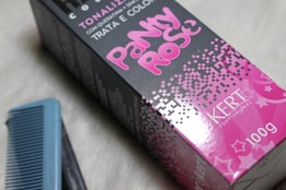 A pink and black hair dye box labeled 'Pinky Rose' with the brand name 'Kert Professional' displayed in white text. The box features pixelated designs and a weight indication of 100g. A blue comb is positioned next to the box on a light-colored surface.