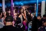 A crowded nightclub scene with people socializing and celebrating. Several individuals are holding bottles with sparklers attached, adding a festive and lively atmosphere. The club has dim lighting with colorful lights enhancing the party ambiance.