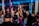 A crowded nightclub scene with people socializing and celebrating. Several individuals are holding bottles with sparklers attached, adding a festive and lively atmosphere. The club has dim lighting with colorful lights enhancing the party ambiance.