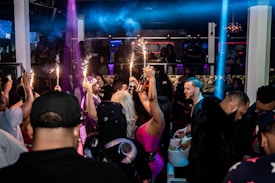 A crowded nightclub scene with people socializing and celebrating. Several individuals are holding bottles with sparklers attached, adding a festive and lively atmosphere. The club has dim lighting with colorful lights enhancing the party ambiance.