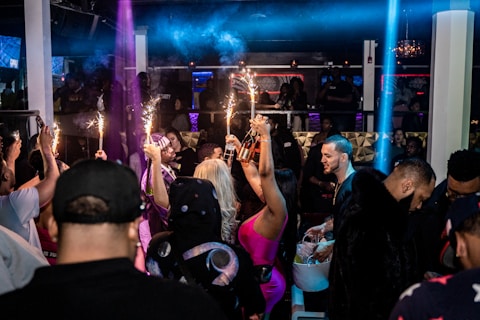A crowded nightclub scene with people socializing and celebrating. Several individuals are holding bottles with sparklers attached, adding a festive and lively atmosphere. The club has dim lighting with colorful lights enhancing the party ambiance.