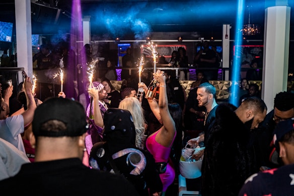 A crowded nightclub scene with people socializing and celebrating. Several individuals are holding bottles with sparklers attached, adding a festive and lively atmosphere. The club has dim lighting with colorful lights enhancing the party ambiance.