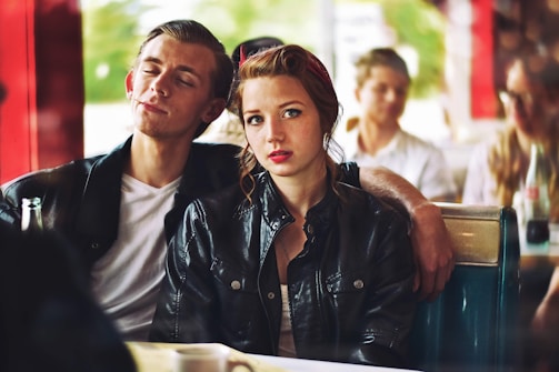 Two young adults are seated in a retro-style diner. The man, with his eyes closed and a relaxed expression, leans on the woman who is looking directly at the camera with a neutral expression. Both are wearing leather jackets. Other patrons are visible in the background, adding to the busy diner atmosphere.