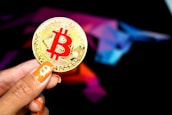 A hand holding a large gold coin with a prominent red Bitcoin symbol. The person's fingernails are painted orange, featuring small Bitcoin icons. In the background, there is a colorful, abstract design resembling a bull, using shades of purple, blue, pink, and black.