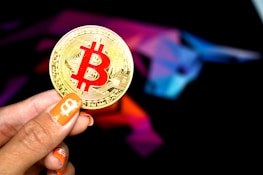 A hand holding a large gold coin with a prominent red Bitcoin symbol. The person's fingernails are painted orange, featuring small Bitcoin icons. In the background, there is a colorful, abstract design resembling a bull, using shades of purple, blue, pink, and black.