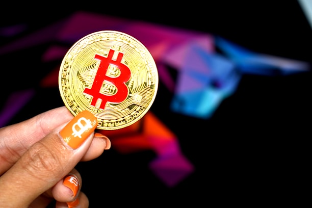 A hand holding a large gold coin with a prominent red Bitcoin symbol. The person's fingernails are painted orange, featuring small Bitcoin icons. In the background, there is a colorful, abstract design resembling a bull, using shades of purple, blue, pink, and black.