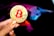 A hand holding a large gold coin with a prominent red Bitcoin symbol. The person's fingernails are painted orange, featuring small Bitcoin icons. In the background, there is a colorful, abstract design resembling a bull, using shades of purple, blue, pink, and black.