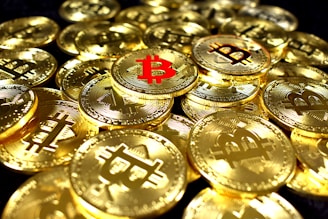 A collection of shiny gold coins featuring the Bitcoin symbol, with one prominent coin having a red Bitcoin symbol. The coins are stacked and scattered, reflecting light with a metallic sheen.