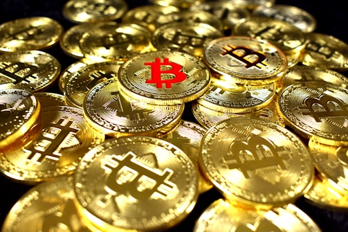 A collection of shiny gold coins featuring the Bitcoin symbol, with one prominent coin having a red Bitcoin symbol. The coins are stacked and scattered, reflecting light with a metallic sheen.