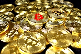 A collection of shiny gold coins featuring the Bitcoin symbol, with one prominent coin having a red Bitcoin symbol. The coins are stacked and scattered, reflecting light with a metallic sheen.