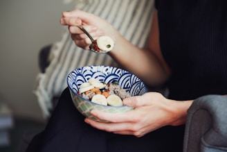 A person's hands holding a bowl of food, with one hand lifting a spoonful towards their mouth. The bowl contains an assortment of items including sliced bananas, nuts, and possibly some grains or cereal. The person is sitting on a couch with a striped pillow in the background.