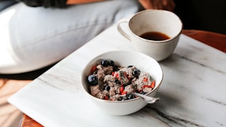 A bowl containing a mixture of chia pudding with goji berries and blueberries is placed on a marble surface. Beside it, a cup filled with black coffee sits. Part of a person's leg and hand are visible in the background, suggesting a relaxed dining or breakfast setting.