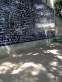 A wall covered in dark blue tiles features the phrase 'I love you' written in multiple languages, each in white text. Small red heart shapes are scattered among the words. The background includes a stone wall with foliage casting shadows on the ground and part of the wall.