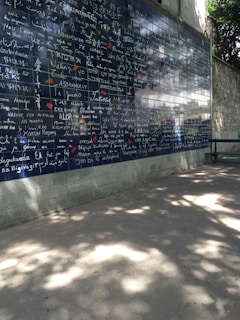 A wall covered in dark blue tiles features the phrase 'I love you' written in multiple languages, each in white text. Small red heart shapes are scattered among the words. The background includes a stone wall with foliage casting shadows on the ground and part of the wall.