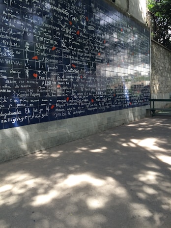 A wall covered in dark blue tiles features the phrase 'I love you' written in multiple languages, each in white text. Small red heart shapes are scattered among the words. The background includes a stone wall with foliage casting shadows on the ground and part of the wall.