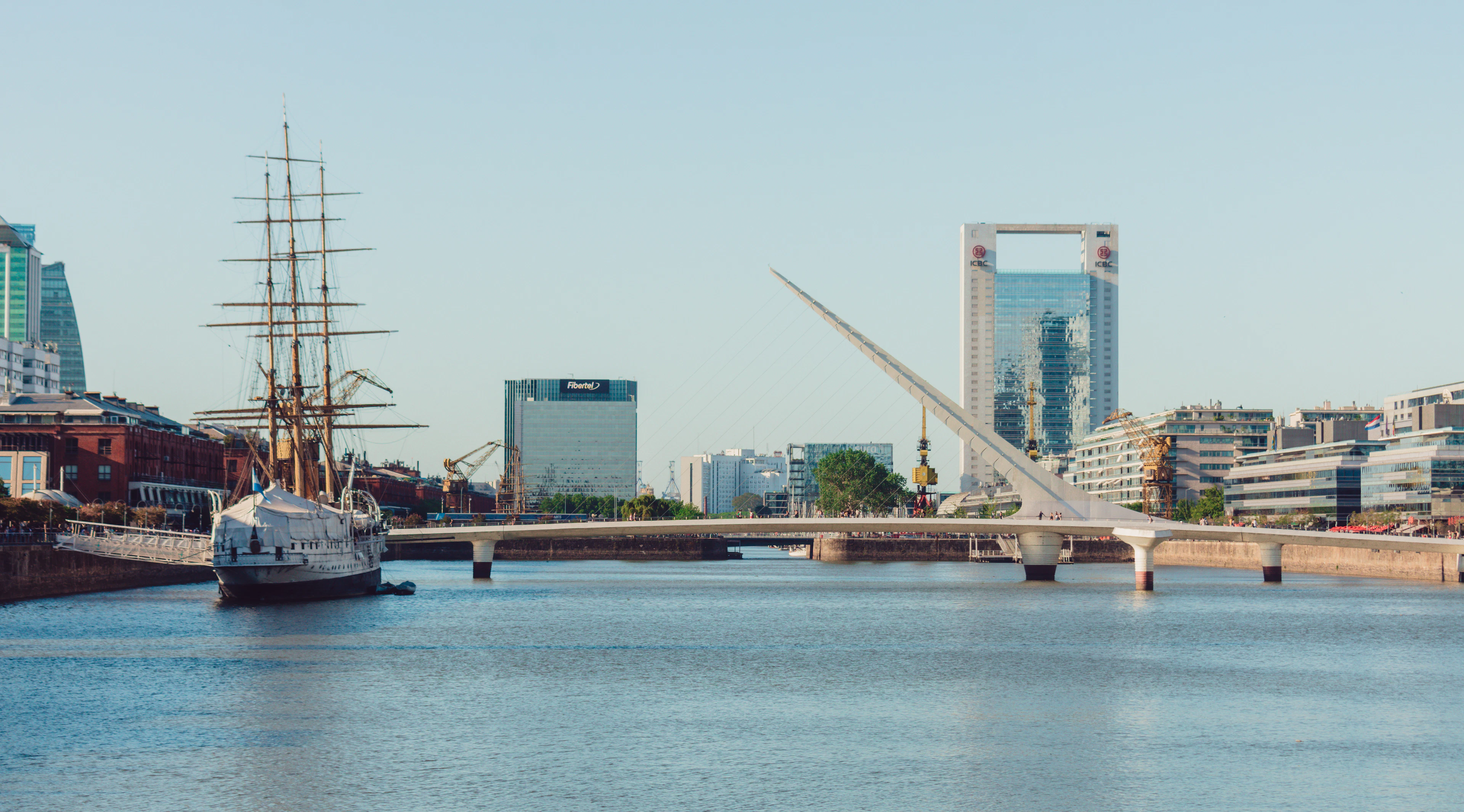 Late afternoon light on the Puerto Madero dock in Buenos Aires, a historic tall ship moored along the old brick quay and the white angled sweep of the Puente de la Mujer set against the district's glass towers.