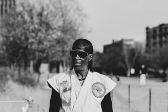 A person wearing a martial arts uniform with patches stands outdoors. They have sunglasses on and the background is slightly out of focus with trees and tall buildings visible.