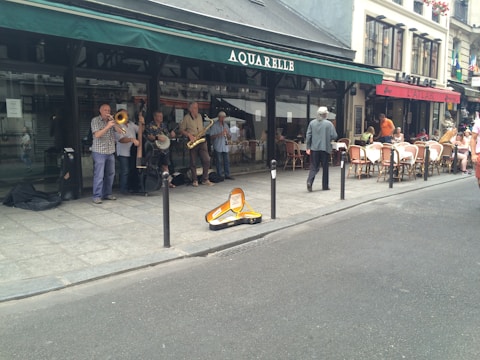 A group of street musicians is performing on a paved sidewalk outside a café. Several musicians are visible with instruments like a trumpet, trombone, banjo, saxophone, and double bass. The setting is urban with a café featuring outdoor seating and people seated at tables. A trombone case is open on the ground, likely for collecting donations.