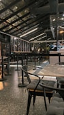 A modern, industrial-style cafe with a glass roof and sleek black furnishings. The space features tables and chairs with cushioned seats and a speckled flooring. Overhead lighting provides a warm ambiance, and bar seating is visible alongside a counter.