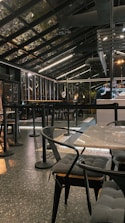 A modern, industrial-style cafe with a glass roof and sleek black furnishings. The space features tables and chairs with cushioned seats and a speckled flooring. Overhead lighting provides a warm ambiance, and bar seating is visible alongside a counter.