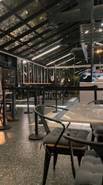 A modern, industrial-style cafe with a glass roof and sleek black furnishings. The space features tables and chairs with cushioned seats and a speckled flooring. Overhead lighting provides a warm ambiance, and bar seating is visible alongside a counter.