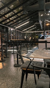 A modern, industrial-style cafe with a glass roof and sleek black furnishings. The space features tables and chairs with cushioned seats and a speckled flooring. Overhead lighting provides a warm ambiance, and bar seating is visible alongside a counter.