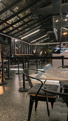 A modern, industrial-style cafe with a glass roof and sleek black furnishings. The space features tables and chairs with cushioned seats and a speckled flooring. Overhead lighting provides a warm ambiance, and bar seating is visible alongside a counter.