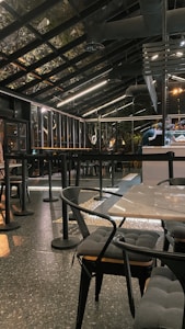 A modern, industrial-style cafe with a glass roof and sleek black furnishings. The space features tables and chairs with cushioned seats and a speckled flooring. Overhead lighting provides a warm ambiance, and bar seating is visible alongside a counter.