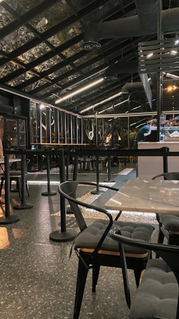 A modern, industrial-style cafe with a glass roof and sleek black furnishings. The space features tables and chairs with cushioned seats and a speckled flooring. Overhead lighting provides a warm ambiance, and bar seating is visible alongside a counter.
