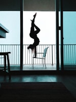 A person is performing an upside-down aerial yoga pose, silhouetted against a bright, light blue sky. The setting is within an open doorway leading to a balcony, with metal railings visible in the background. The interior appears dark, with minimal light filtering through.