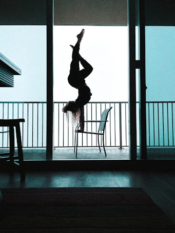 A person is performing an upside-down aerial yoga pose, silhouetted against a bright, light blue sky. The setting is within an open doorway leading to a balcony, with metal railings visible in the background. The interior appears dark, with minimal light filtering through.