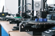 Several 3D printers are lined up on a wooden table, each actively engaged in printing blue cylindrical objects. The machines appear modern, with visible wiring and components, and the work environment suggests a focus on technology and innovation.