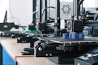 Several 3D printers are lined up on a wooden table, each actively engaged in printing blue cylindrical objects. The machines appear modern, with visible wiring and components, and the work environment suggests a focus on technology and innovation.