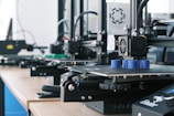 Several 3D printers are lined up on a wooden table, each actively engaged in printing blue cylindrical objects. The machines appear modern, with visible wiring and components, and the work environment suggests a focus on technology and innovation.