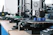 Several 3D printers are lined up on a wooden table, each actively engaged in printing blue cylindrical objects. The machines appear modern, with visible wiring and components, and the work environment suggests a focus on technology and innovation.