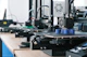 Several 3D printers are lined up on a wooden table, each actively engaged in printing blue cylindrical objects. The machines appear modern, with visible wiring and components, and the work environment suggests a focus on technology and innovation.