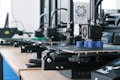 Several 3D printers are lined up on a wooden table, each actively engaged in printing blue cylindrical objects. The machines appear modern, with visible wiring and components, and the work environment suggests a focus on technology and innovation.