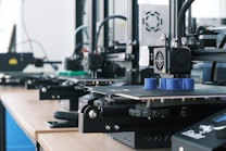 Several 3D printers are lined up on a wooden table, each actively engaged in printing blue cylindrical objects. The machines appear modern, with visible wiring and components, and the work environment suggests a focus on technology and innovation.