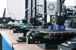 Several 3D printers are lined up on a wooden table, each actively engaged in printing blue cylindrical objects. The machines appear modern, with visible wiring and components, and the work environment suggests a focus on technology and innovation.