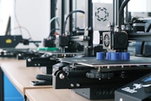 Several 3D printers are lined up on a wooden table, each actively engaged in printing blue cylindrical objects. The machines appear modern, with visible wiring and components, and the work environment suggests a focus on technology and innovation.