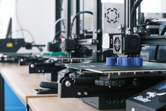 Several 3D printers are lined up on a wooden table, each actively engaged in printing blue cylindrical objects. The machines appear modern, with visible wiring and components, and the work environment suggests a focus on technology and innovation.