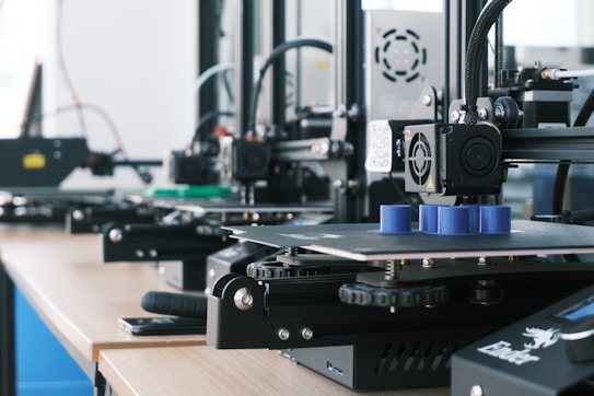 Several 3D printers are lined up on a wooden table, each actively engaged in printing blue cylindrical objects. The machines appear modern, with visible wiring and components, and the work environment suggests a focus on technology and innovation.