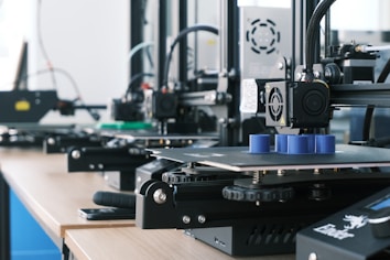 Several 3D printers are lined up on a wooden table, each actively engaged in printing blue cylindrical objects. The machines appear modern, with visible wiring and components, and the work environment suggests a focus on technology and innovation.