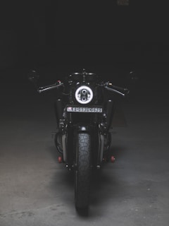 A motorcycle is positioned directly facing the viewer, illuminated by a focused light that highlights its front features. The scene is set in a dimly lit environment, emphasizing the bike's sleek black and metallic design. The license plate is visible, and the surroundings appear to be an empty, dark space, possibly indoors.
