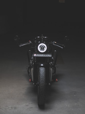 A motorcycle is positioned directly facing the viewer, illuminated by a focused light that highlights its front features. The scene is set in a dimly lit environment, emphasizing the bike's sleek black and metallic design. The license plate is visible, and the surroundings appear to be an empty, dark space, possibly indoors.