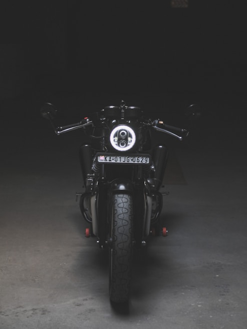 A motorcycle is positioned directly facing the viewer, illuminated by a focused light that highlights its front features. The scene is set in a dimly lit environment, emphasizing the bike's sleek black and metallic design. The license plate is visible, and the surroundings appear to be an empty, dark space, possibly indoors.