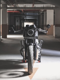 A motorcycle is positioned in a dimly lit parking garage, viewed directly from the front. The space has an industrial feel with concrete walls and a network of pipes on the ceiling. The focus on the motorcycle’s headlight and handlebars adds to the dramatic composition.