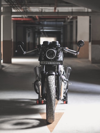 A motorcycle is positioned in a dimly lit parking garage, viewed directly from the front. The space has an industrial feel with concrete walls and a network of pipes on the ceiling. The focus on the motorcycle’s headlight and handlebars adds to the dramatic composition.